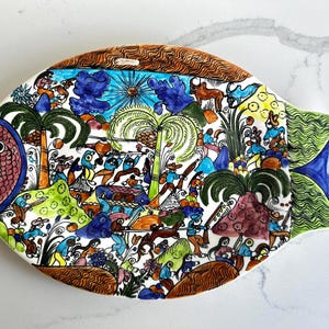 May include: A colorful, hand-painted ceramic fish-shaped platter. The design features a detailed scene with figures, palm trees, and various elements in shades of blue, green, orange, and brown. The fish has a red and blue eye and tail.
