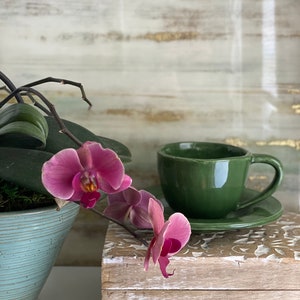 May include: A green teacup and saucer set on a wooden box with a carved design. A pink orchid with green leaves is in the foreground.