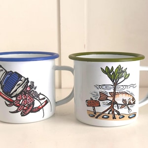 Puede incluir: Dos tazas de esmalte blanco con ilustraciones. Una presenta un cangrejo ermitaño rojo y azul, la otra una escena de peces y manglares. Ambas tazas tienen un borde y un asa de color.