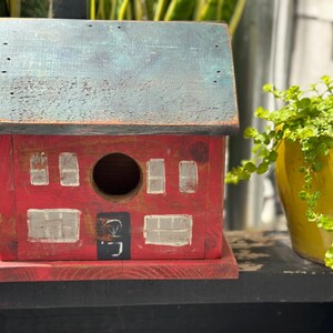 Può includere: Una casetta per uccelli rustica in legno rosso con finitura anticata. La casetta per uccelli presenta un tetto blu-grigio, un foro di ingresso circolare e ritagli di finestre rettangolari. Una piccola porta nera è dipinta sul davanti. Un'aggiunta affascinante a qualsiasi giardino.