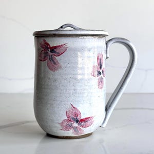 Puede incluir: Una taza de cerámica hecha a mano con tapa, con un esmalte blanco moteado y diseños florales rojos y negros pintados a mano. La taza tiene un asa curva y una tapa a juego. La taza mide aproximadamente 13 cm de altura.