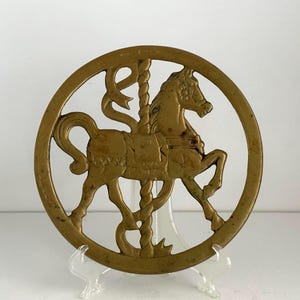 Op de afbeelding: Een vintage messing carrousel paard ornament. Het paard staat in een cirkelvormig frame met een decoratieve paal en linten. Het paard staat in een drafhouding, met uitgestrekte benen. Het messing heeft een warme, verouderde patina, waardoor het een klassieke uitstraling heeft.