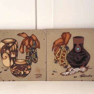 May include: Two square ceramic tiles with Southwestern-style designs. One tile features two pottery vessels and corn. The other tile shows a dark brown vase, corn, and a feather. The tiles have a beige background.