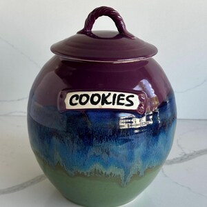 May include: A round ceramic cookie jar with a glossy finish, featuring a gradient of purple at the top, transitioning into deep blue, and ending in a light green base. It has a matching lid with a twisted handle and a label reading COOKIES.