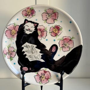 May include: A white ceramic plate with a black and white cat painted on it. The cat is surrounded by pink flowers with yellow centers. The plate is on a black metal stand.