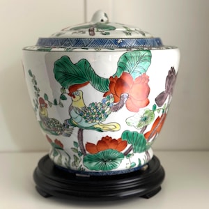 Puede incluir: Un jarrón de cerámica blanco con tapa, con un diseño floral y de aves colorido. El jarrón tiene una base de madera negra. El diseño incluye detalles en rojo, naranja, verde y azul. Es un objeto decorativo.