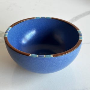 Peut inclure: Un petit bol rond en céramique bleue avec un bord marron et une bande décorative de carrés bleus et blancs. L'intérieur du bol est lisse et brillant. Le bol est posé sur un fond blanc.
