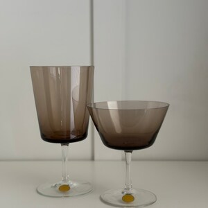 May include: Two clear glass drinking glasses with a brown tint. The larger glass is a wine glass and the smaller glass is a champagne flute. Both glasses have a gold sticker on the bottom.