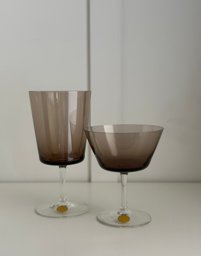 May include: Two clear glass drinking glasses with a brown tint. The larger glass is a wine glass and the smaller glass is a champagne flute. Both glasses have a gold sticker on the bottom.