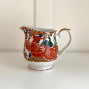 Puede incluir: Una jarra de leche de porcelana blanca con un diseño pintado a mano de un pájaro naranja grande, un pájaro verde más pequeño y motivos florales en rojo, azul y dorado. La jarra tiene un asa curva, un borde festoneado y una base.