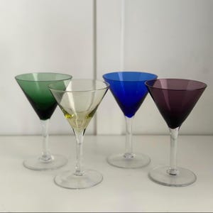 Könnte beinhalten: Vier farbige Martinigläser. Die Gläser haben klare Stiele und Böden, mit Schalen in Grün, Gelb, Blau und Lila. Sie sind auf einer weißen Oberfläche mit weißem Hintergrund angeordnet.