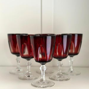 Puede incluir: Juego de seis copas de vino tinto rubí. Cada copa presenta un diseño de cuenco facetado y un tallo transparente y ornamentado. Las copas están dispuestas sobre una superficie blanca, mostrando su elegante forma y rico color.