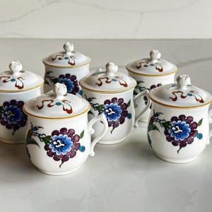 Puede incluir: Seis tazas de té de cerámica blanca con tapas. Cada taza presenta un diseño floral con detalles en azul, morado y verde. Las tapas tienen un borde decorativo y un pequeño pomo. Las tazas tienen asas pequeñas.