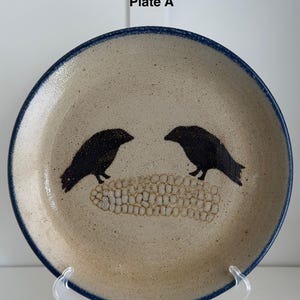 Puede incluir: Un plato de cerámica con un borde azul presenta dos siluetas negras de pájaros posados ​​en una mazorca de maíz. El plato está etiquetado como "Plate A".