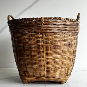 May include: A tall, round, dark brown woven wicker basket featuring a tight, horizontal weave pattern, a reinforced vertical spine, and two small, integrated handles on the upper rim. The basket has a sturdy, flat base and a wide, open top.