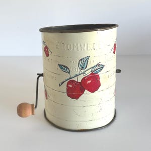 以下が含まれることがあります： クリーム色のヴィンテージキッチンふるい。手回しと木製のハンドルが付いています。赤いサクランボのイラストと「Cromwell」のエンボス加工が施されています。側面に測定マークが見えます。