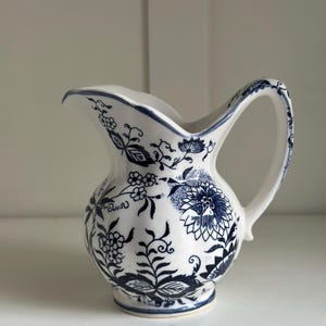May include: A small white ceramic pitcher with a blue floral pattern. The pitcher has a rounded body and a curved handle.
