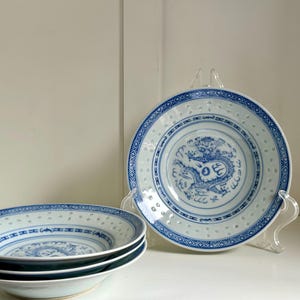 Op de afbeelding: Een set witte en blauwe porseleinen kommen met een drakenmotief. De kommen hebben een geperforeerde rand en een blauwe draak in het midden. Eén kom wordt op een transparante standaard getoond.