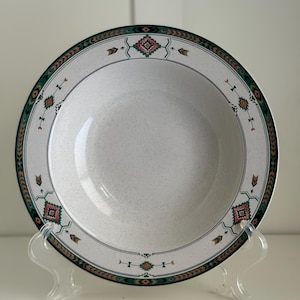 Op de afbeelding: Een witte keramische soepkom met een groen, bruin en roze geometrisch patroon rond de rand. De kom staat op een transparante kunststof standaard.