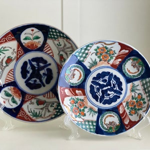 Op de afbeelding: Twee decoratieve keramische borden met een blauw-wit ontwerp. De borden hebben een bloemenpatroon, geometrische vormen en een gestileerde vogel. De borden staan op presentatiestandaards.