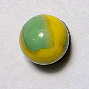 Op de afbeelding: Een bolvormige glazen knikker met een glad, gepolijst oppervlak en een wervelend patroon van ondoorzichtig felgeel en zacht saliegroen. De knikker heeft een glanzende afwerking die het licht reflecteert en een kleine witte lichtvlek creëert.