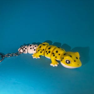 Könnte beinhalten: Ein gelb-weißer, beweglicher Gecko-Schlüsselanhänger mit schwarzen Punkten auf dem Körper. Die Echsenfigur hat eine glatte Oberfläche, große dunkle Augen und ist mit einem kurzen Kettenglied an einem silberfarbenen Metallring befestigt.