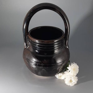 May include: A black ceramic vase with a wide, round body and a handle. The vase has a glossy finish and is decorated with subtle brown lines. White flowers are placed in front of the vase.