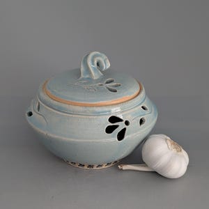May include: A light blue ceramic garlic keeper with a lid featuring a decorative handle and floral cutouts. The keeper has a light brown rim and a matching floral design around the base. A whole garlic bulb rests beside the keeper.