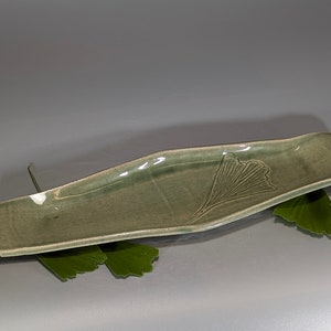 May include: A celadon green ceramic serving dish with a leaf design. The rectangular dish has gently curved sides and a raised leaf pattern on one side. The dish is approximately 12 inches long.