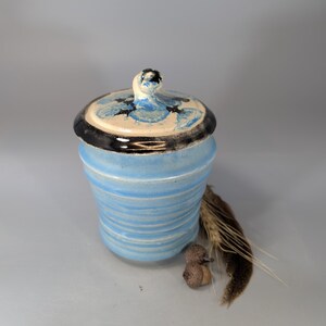 May include: A handmade ceramic jar with a lid. The jar is light blue with horizontal ridges and a black rim. The lid has a white base with blue and black accents. A decorative piece.