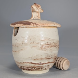 May include: A handmade ceramic honey pot with a lid and a wooden honey dipper. The pot is off-white with brown accents and a decorative pattern at the base. The lid has a unique handle. A functional and decorative kitchen item.