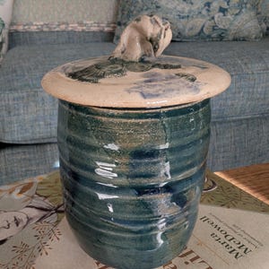 May include: A handcrafted ceramic jar with a lid. The jar is a deep teal color with horizontal ridges. The lid is off-white with blue and green painted details and a sculpted handle. The jar is sitting on a book.