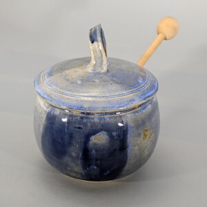 May include: A handmade ceramic honey pot with a lid and a wooden honey dipper. The pot is glazed in shades of blue and tan, with a textured surface. The lid has a curved handle.