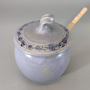 May include: A light blue ceramic jar with a lid and a wooden spoon. The jar has a rounded shape with a lid featuring a decorative floral border. The lid has a curved handle. The wooden spoon rests on the side of the jar.