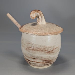 May include: A handmade ceramic jar with a lid and a wooden spoon. The jar is off-white with brown swirl patterns and a decorative band at the base. The lid has a unique, curved handle. The spoon rests on the lid.