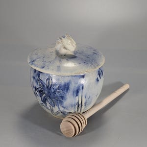 May include: A ceramic honey pot with a lid, featuring a blue floral design and a wooden honey dipper. The pot is round with a speckled blue and white glaze. The honey dipper is light-colored wood.