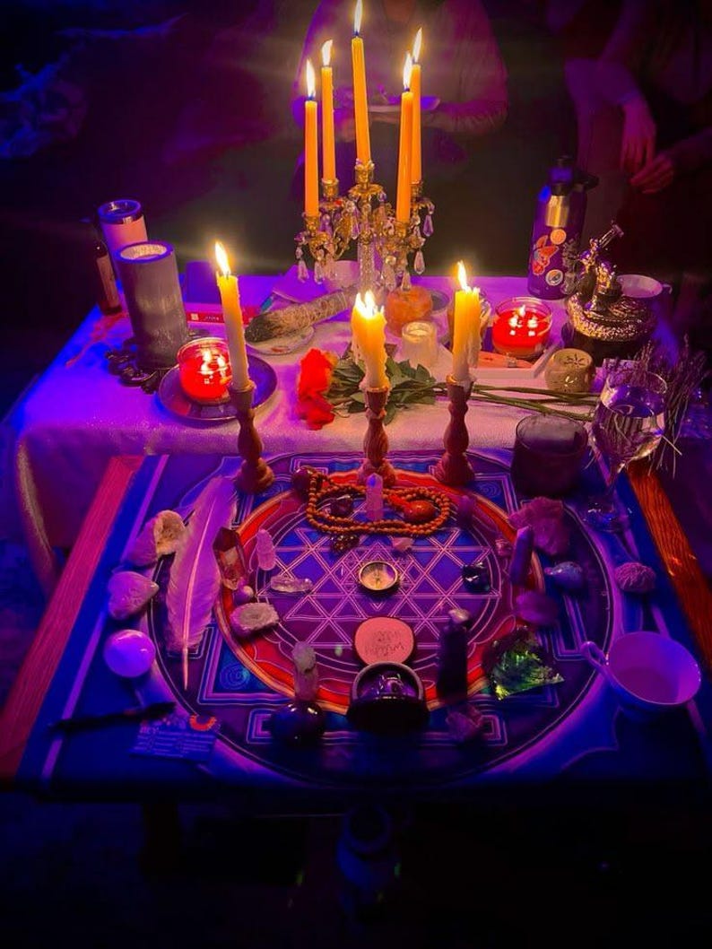 May include: A ritual altar setup featuring a central blue and purple circular cloth with a geometric star pattern. The arrangement includes multiple lit yellow taper candles in holders, various crystals, a white feather, a compass, and beads.