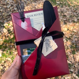 Puede incluir: Un sobre de papel rojo oscuro que contiene una tarjeta de reseña de libro, una pluma negra y una vela pequeña. Una cinta de terciopelo negro ata el paquete, con una etiqueta de un cuervo negro y un marcapáginas rojo.