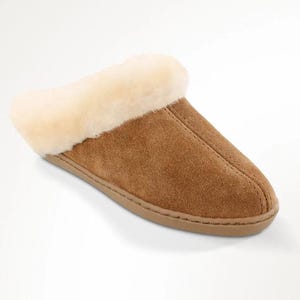 May include: A tan suede slipper featuring a round toe, a center seam detail, and a thick, cream-colored plush lining that extends over the collar. The slipper has a flat, tonal sole and a slip-on design with an open back for easy entry.