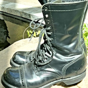 Könnte beinhalten: Ein schwarzer Kampfstiefel aus Leder mit hohem Schaft, verstärkter Zehenkappe mit Ziernähten, Schnürverschluss mit Metallösen und einer dicken, strapazierfähigen Gummisohle. Der Stiefel hat eine glatte, polierte Oberfläche.