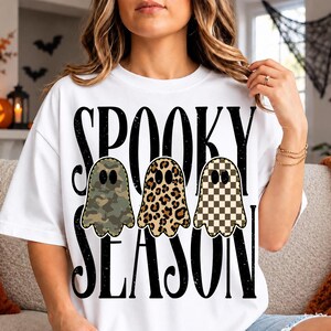 Può includere: Una t-shirt bianca a maniche corte con il testo nero SPOOKY SEASON e tre illustrazioni di fantasmi. Il primo fantasma ha un motivo mimetico verde, il secondo una stampa leopardata marrone e il terzo un motivo a scacchi beige e marrone.