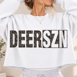 May include: A white long-sleeve cropped sweatshirt featuring the text DEERSZN printed across the chest. The letters are filled with a dark green and brown camouflage pattern. The garment has a crew neckline and a raw, unfinished hemline.