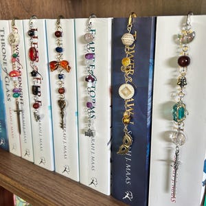 May include: Six metal book markers hanging from the tops of book spines. Each marker features a silver or gold chain adorned with various beads, charms, and pendants, including miniature swords, keys, masks, stars, and books, resting against book covers.