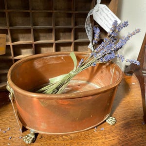May include: An oval copper basin with a polished metallic finish, featuring four ornate brass claw feet and two side handles. A bundle of dried purple lavender stems tied with a light green ribbon rests inside the basin, which has a smooth interior.