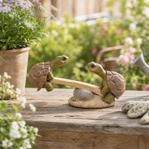 Op de afbeelding: Een decoratief tuinornament met twee kleine bruine en groene schildpadden die aan weerszijden van een houten wip zitten. De wip balanceert op een gestructureerde, steenachtige basis. De schildpadden hebben gedetailleerde schilden en realistische koppen.