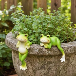 Könnte beinhalten: Zwei hellgrüne Froschfiguren mit großen dunklen Augen und weißen Bäuchen. Ein Frosch steht aufrecht mit den Händen auf dem Rand eines Steinkübels, während der andere Frosch horizontal liegt und seine Beine über den Rand des Kübels hängen.