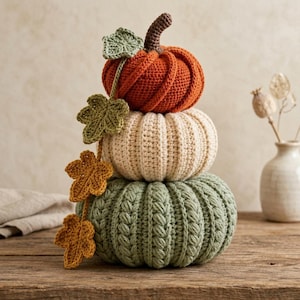 May include: A stacked trio of crocheted pumpkins in orange, cream, and sage green. The top pumpkin has a brown stem. A vine of crocheted leaves in shades of light green, dark green, and mustard yellow hangs down the side of the stacked pumpkins.