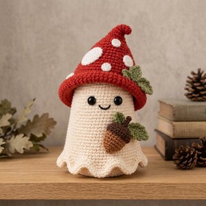 May include: A crocheted mushroom-shaped figure with a cream-colored body and a red cap featuring white spots. The figure has black bead eyes, a stitched smile, and is decorated with a small brown acorn and green crocheted leaves.