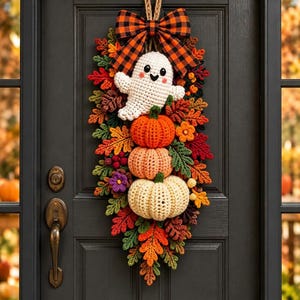 May include: A crochet door hanging featuring a white ghost, three stacked pumpkins in orange, peach, and cream, and colorful autumn leaves. The arrangement is topped with a large orange and black plaid bow and hangs from a braided rope loop.