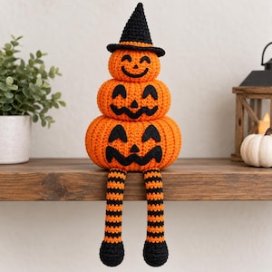 May include: A crocheted stack of three orange pumpkins with black felt jack-o'-lantern faces, topped with a black witch hat. The bottom pumpkin has two long, dangling legs featuring horizontal orange and black stripes with black feet at the ends.